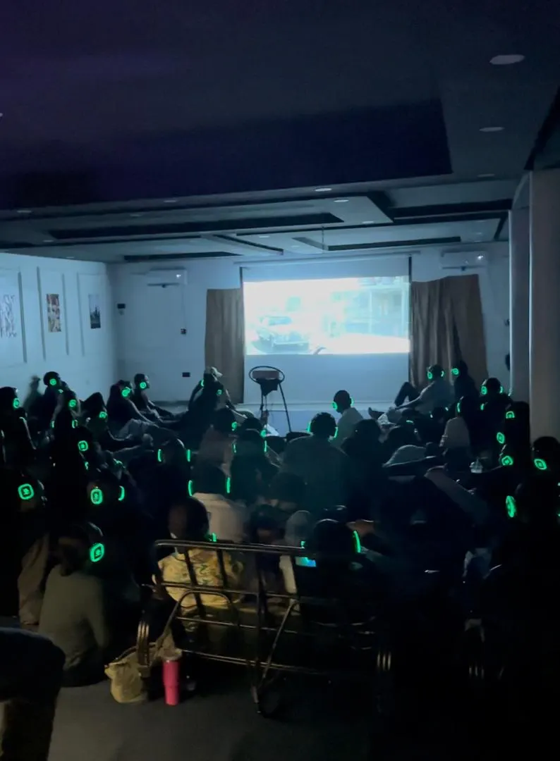 A full room in the dark facing the big screen, everyone wearing glowing green headphones